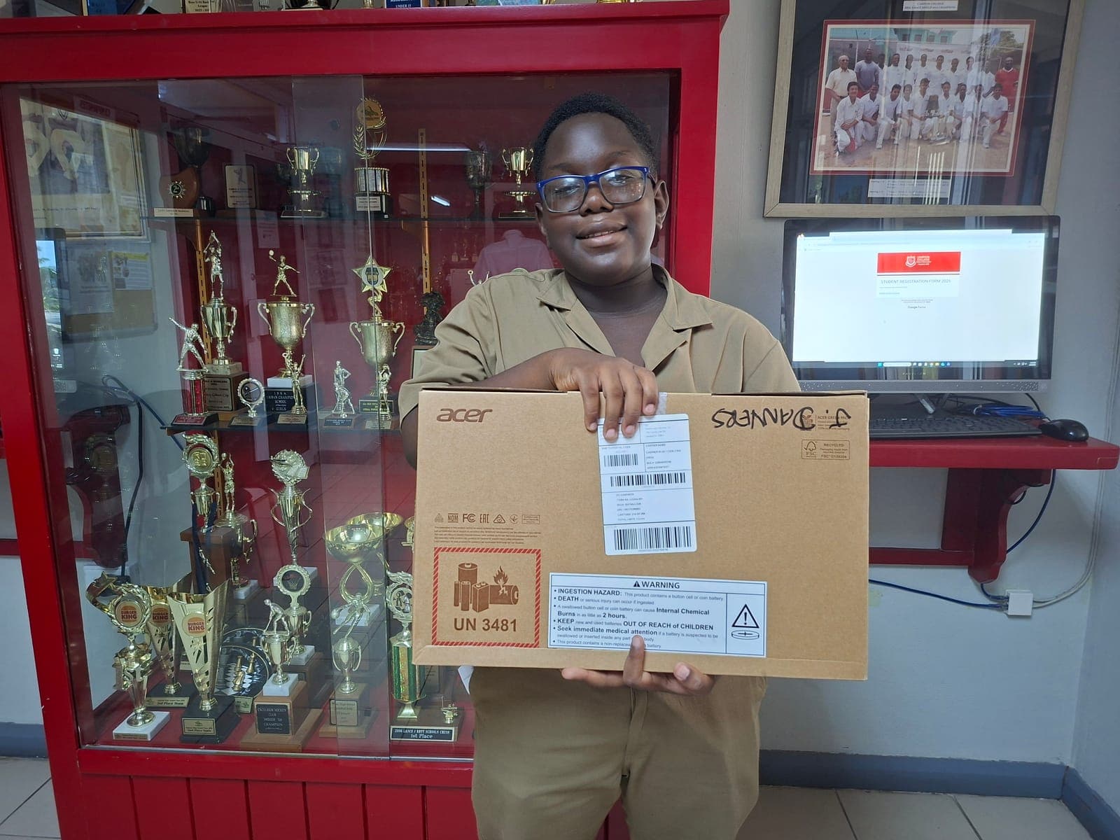 A Campion College student in glasses holding up a new laptop, still boxed, in front of the school’s trophy cabinet.