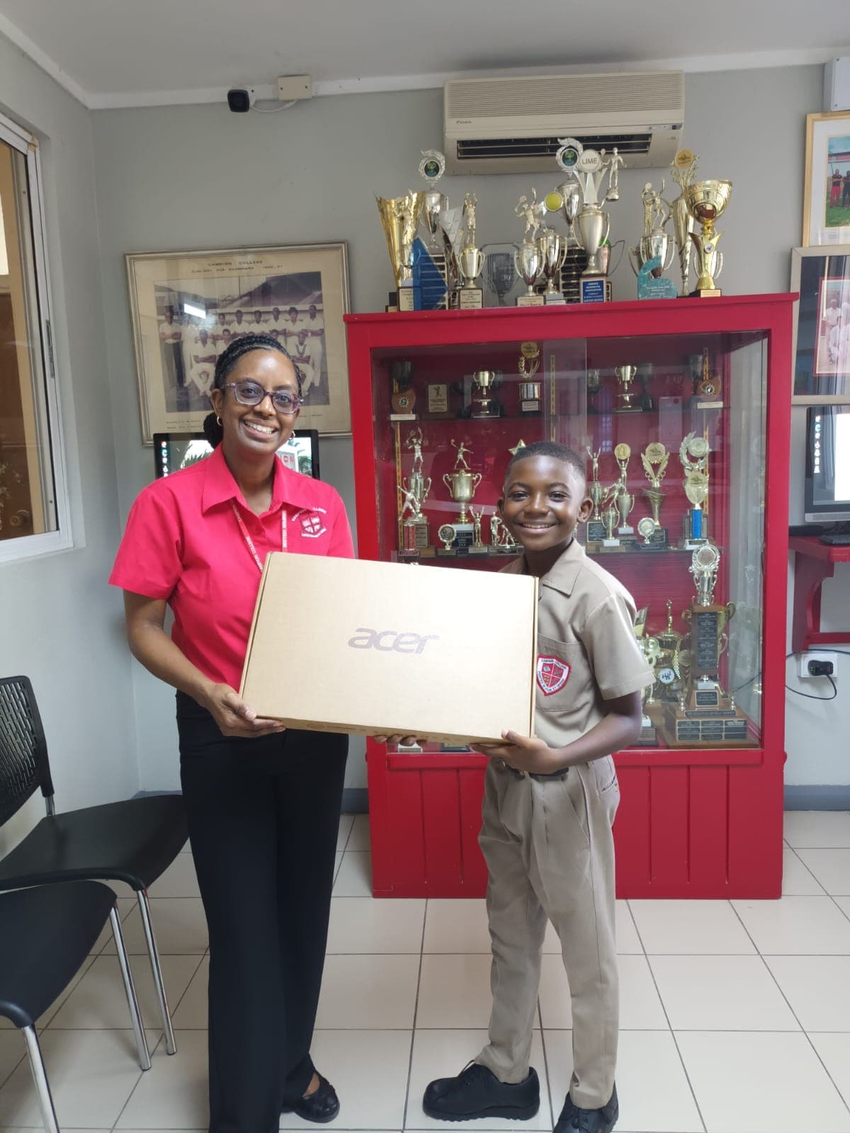 A smiling Campion College student receiving a boxed laptop from a member of staff beside the trophy cabinet.